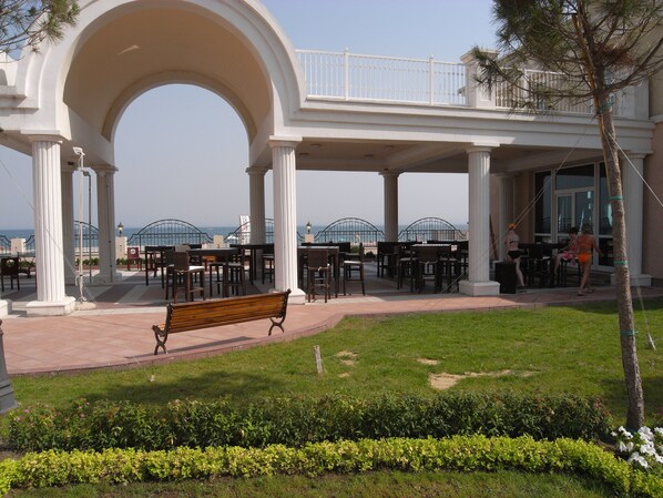 A covered outdoor seating area with tables and chairs, a bench, and a view of the sea. - Sunset Resort (Pomorie)