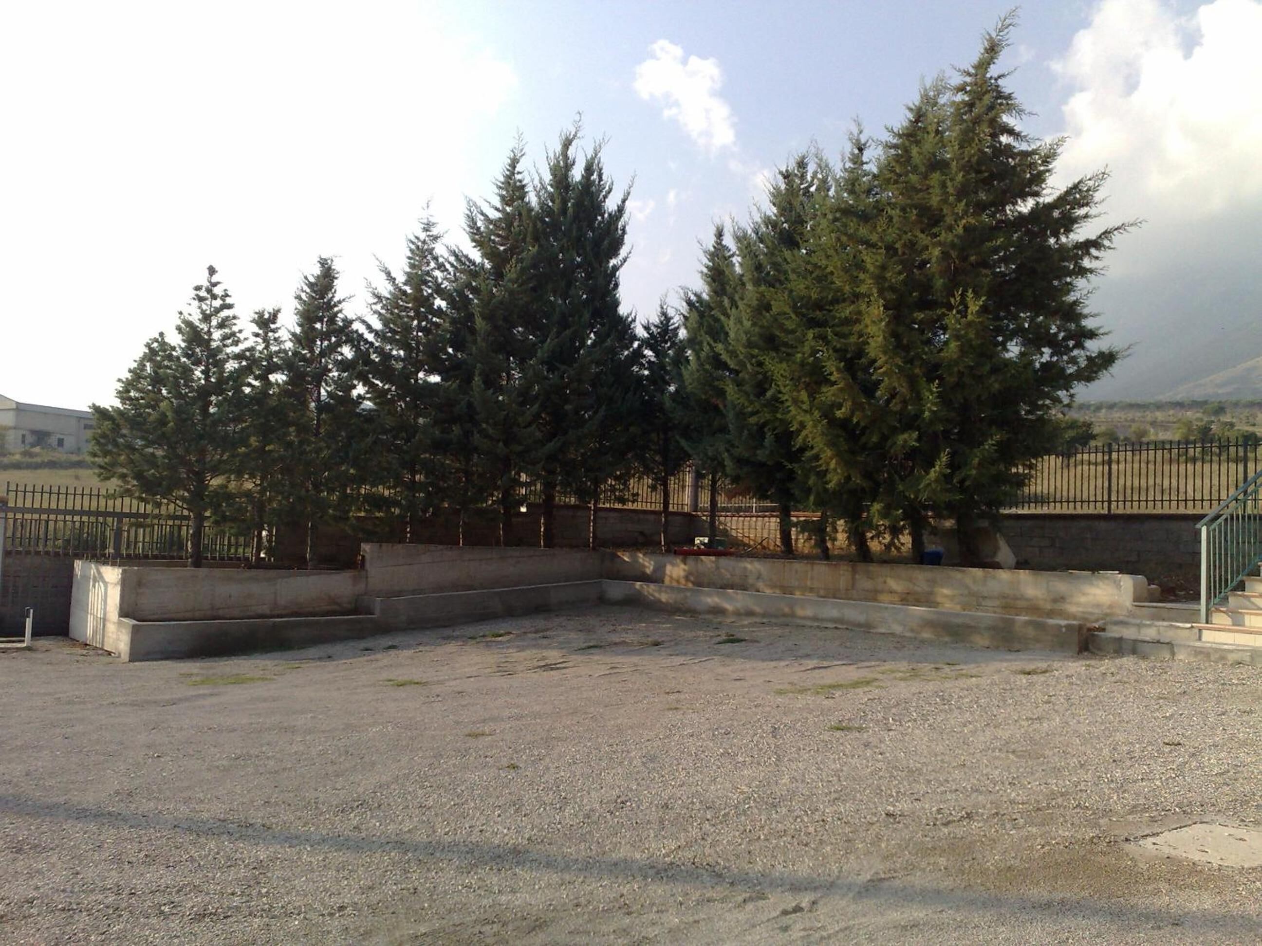 A gravel parking area with a concrete barrier and steps leading to a fenced area with tall evergreen trees.
