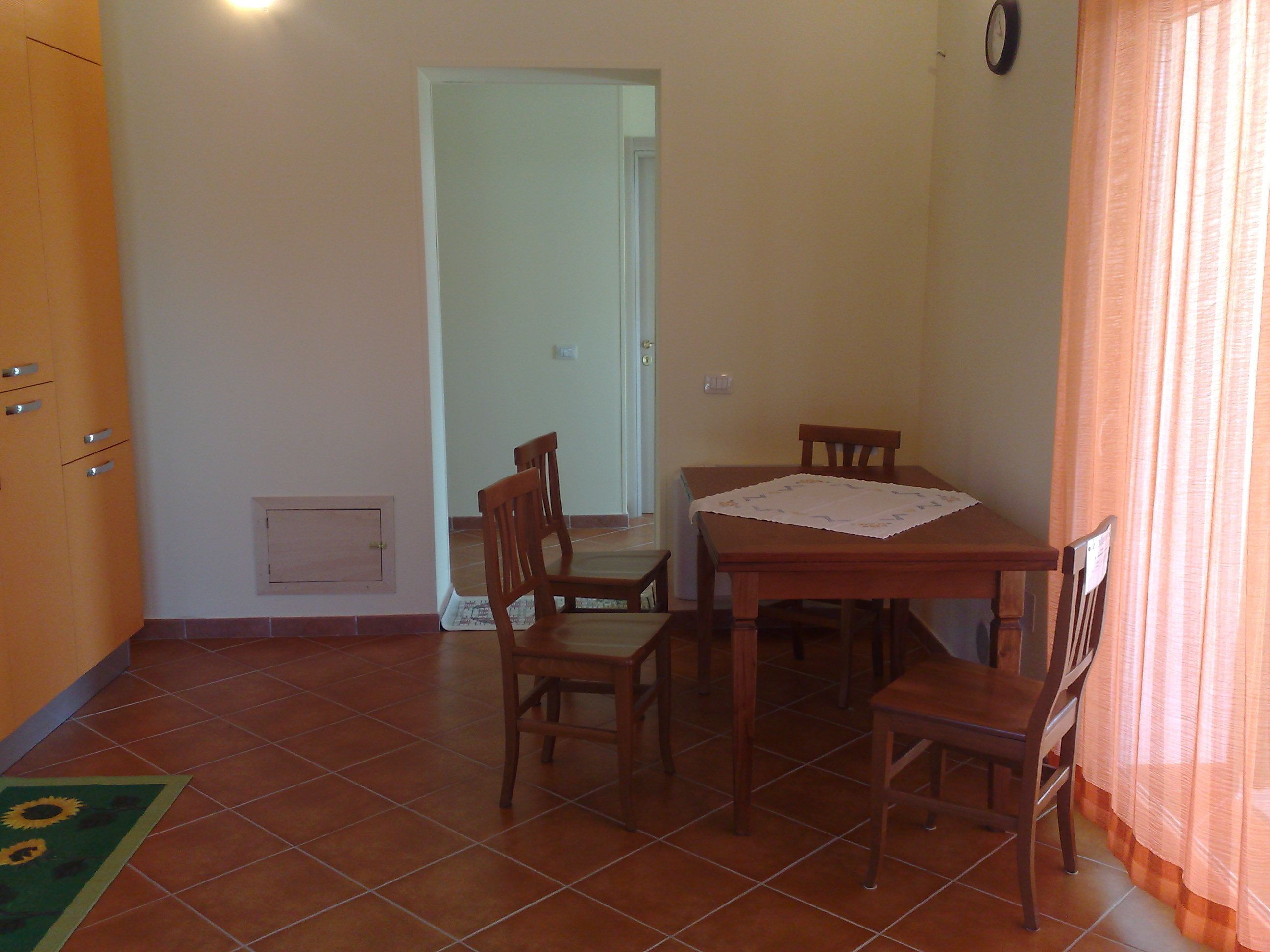 Un coin repas avec une table et des chaises en bois, une horloge au mur et une porte donnant sur une autre pièce.
