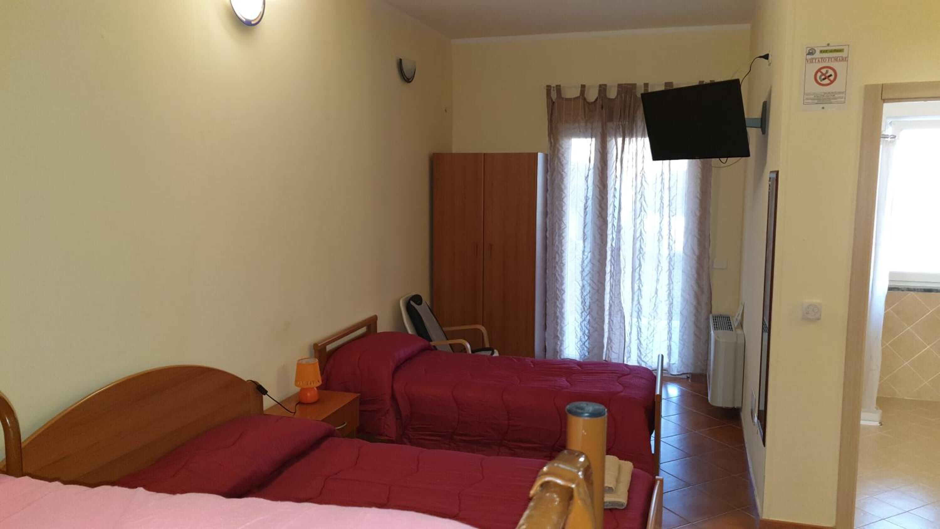 A bedroom with two beds, a wooden wardrobe, a television mounted on the wall, and a window with curtains.