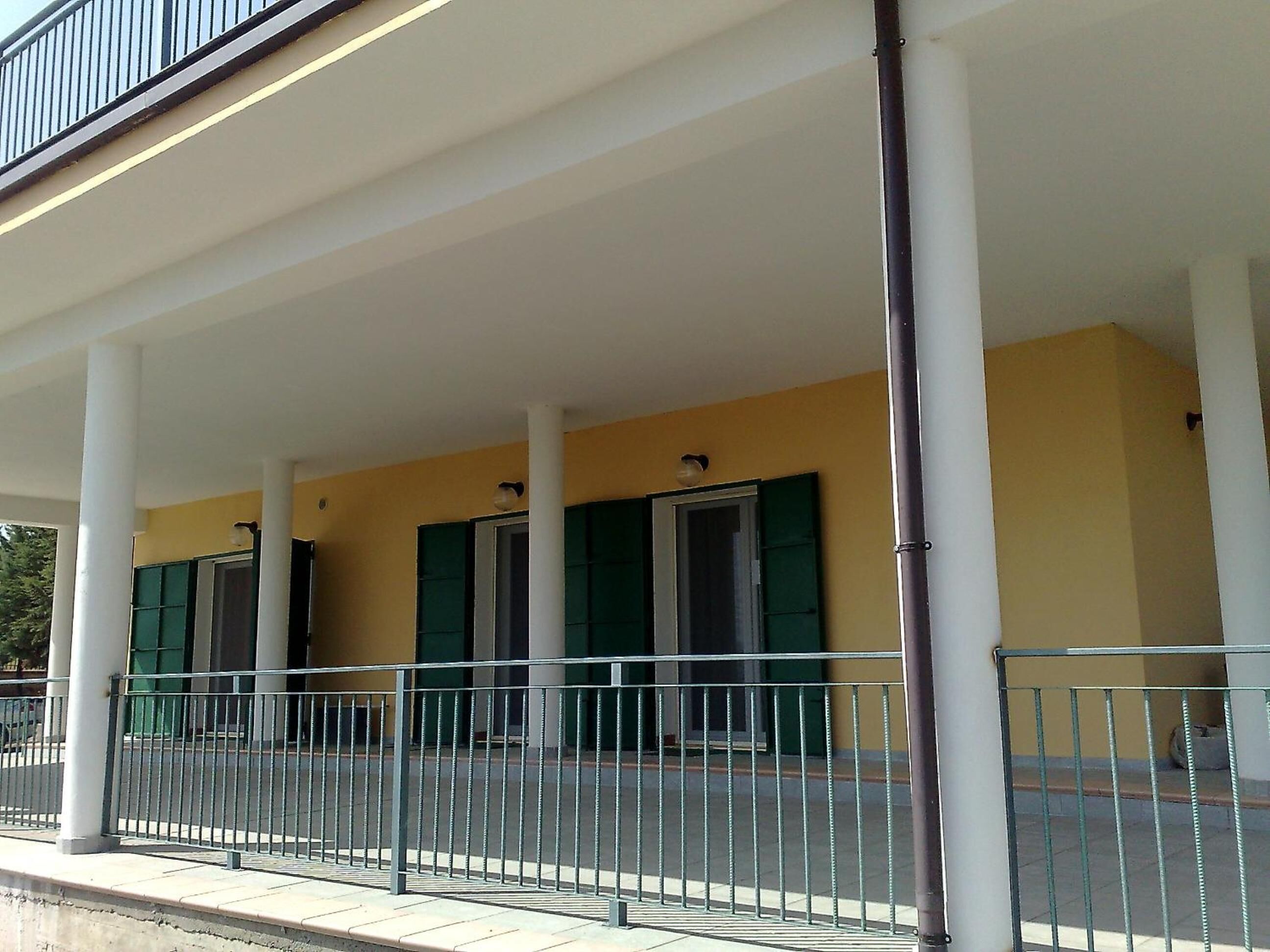 A covered outdoor area with a metal railing, columns, and doors with green shutters.