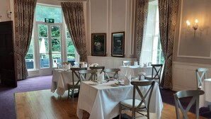 Un comedor con mesas dispuestas para el servicio, ventanas grandes con cortinas y obras enmarcadas en las paredes.