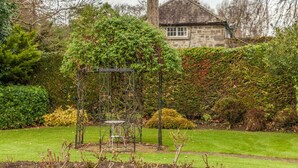 Un jardín con un pórtico de metal, una casa de piedra al fondo y diversos arbustos y árboles.