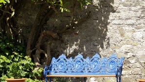 Un banco azul ornamentado con diseños intrincados, situado frente a un muro de piedra con un árbol y plantas en macetas cerca.