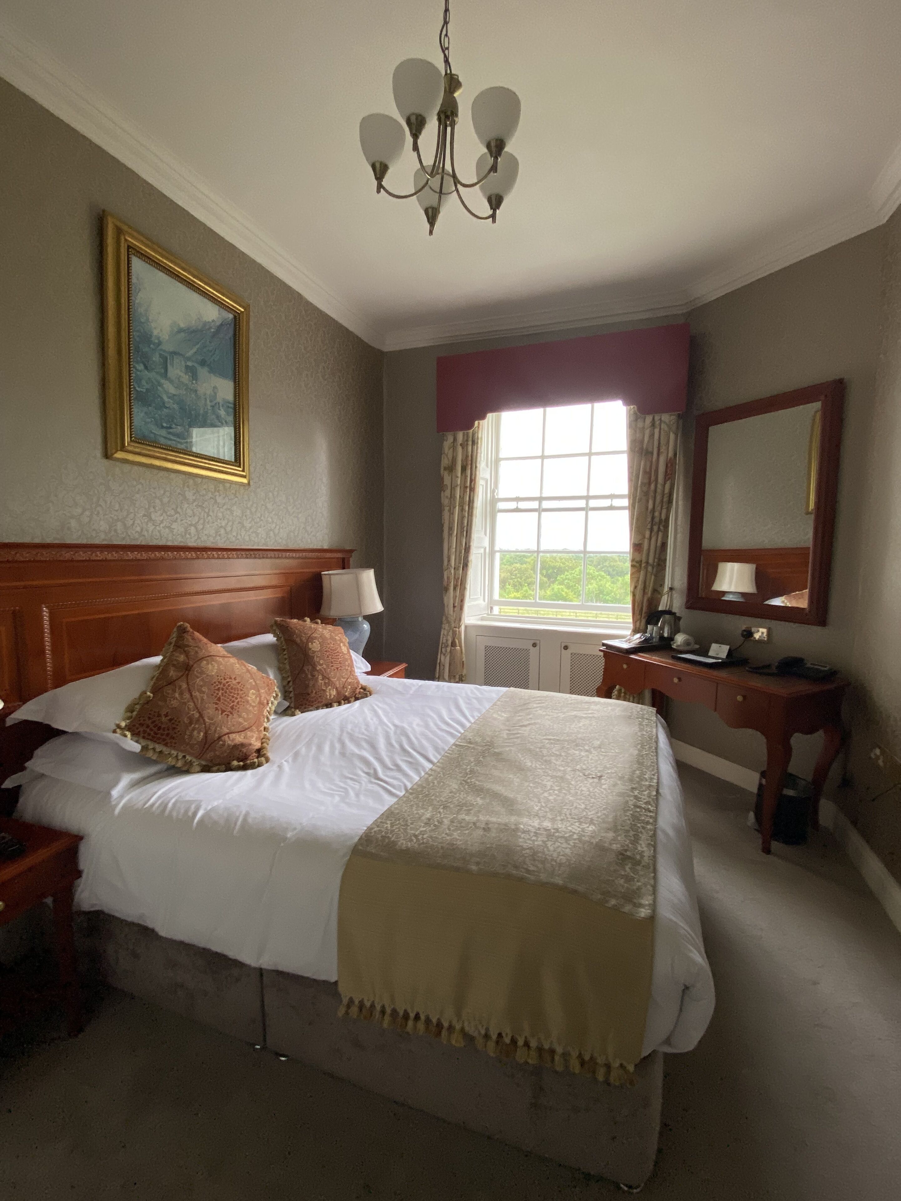 A hotel room with a large bed, a wooden headboard, a desk with a mirror, a window with curtains, and a chandelier.
