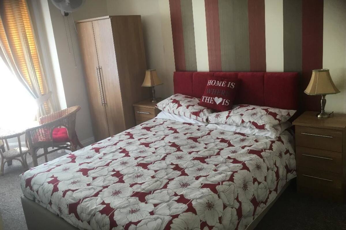 A bedroom with a floral bedspread, a red headboard, two bedside lamps, a wooden wardrobe, and a chair by the window.
