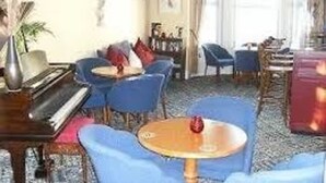 Olohuoneessa on piano, siniset tuolit, pyöreä pöytä ja punainen maljakko.