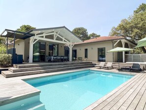 Une piscine avec une terrasse en bois, un patio couvert doté de tables et de chaises, et un petit bâtiment surmonté d’un toit en tuiles.
