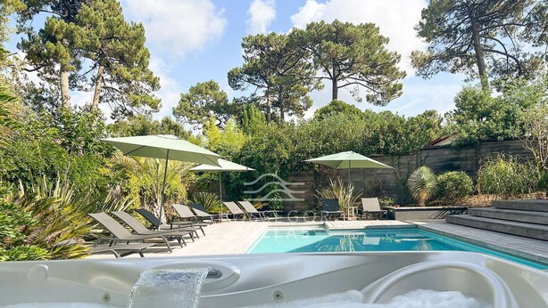 Un bain à remous avec de l’eau en mouvement, entouré de chaises longues et de parasols, avec une piscine et des arbres en arrière-plan.