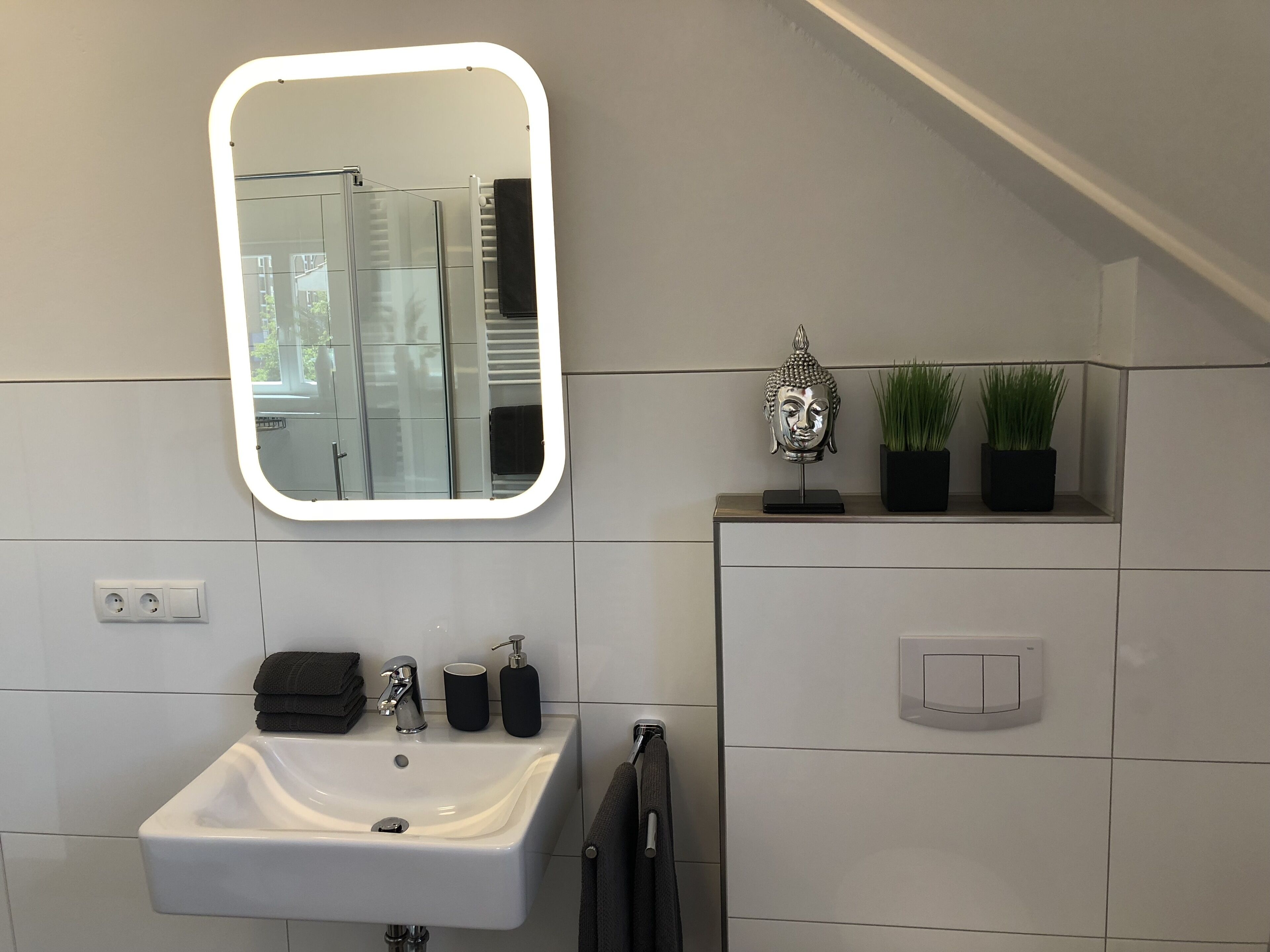 A modern bathroom with a white sink, a mirror with a light, a towel rack, a Buddha head sculpture, and two potted plants.