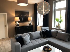 Un salon moderne avec un canapé d’angle gris, deux lampadaires, un meuble noir et une grande fenêtre donnant sur une plante.