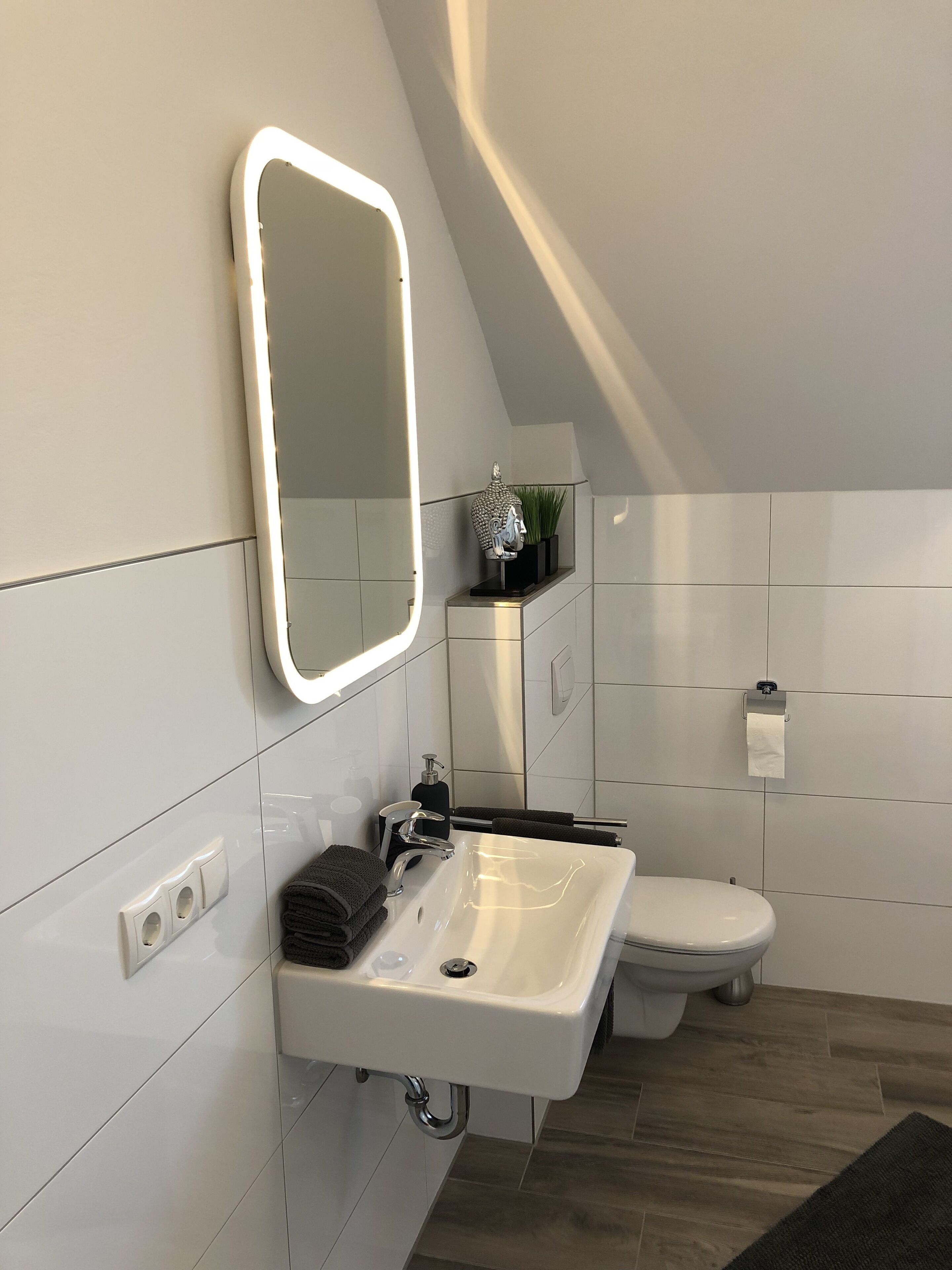 Une salle de bain moderne avec un lavabo blanc, un miroir et des toilettes. On y trouve un porte-serviettes avec des serviettes pliées et une petite plante posée sur le meuble-lavabo.