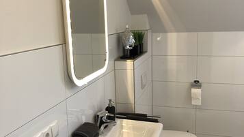 A modern bathroom with a white sink, mirror, and toilet. There is a towel rack with folded towels and a small plant on the vanity.