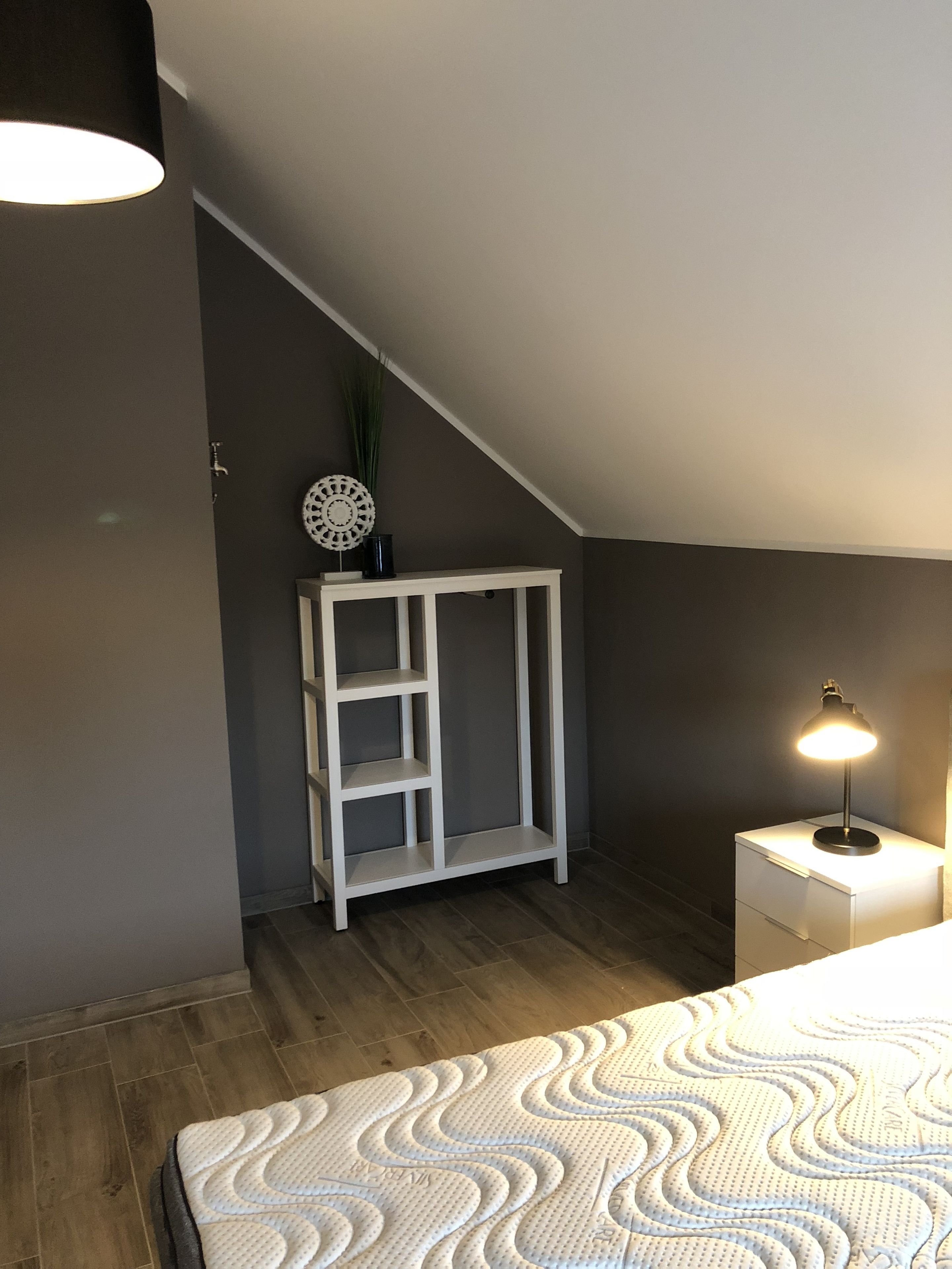 A bedroom with a sloped ceiling, a white bedside table with a lamp, a white shelving unit, and a patterned bedspread.