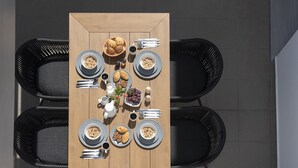 Una mesa rectangular de madera preparada para el desayuno, con platos de cereales, cruasanes y fruta, rodeada de sillas negras.