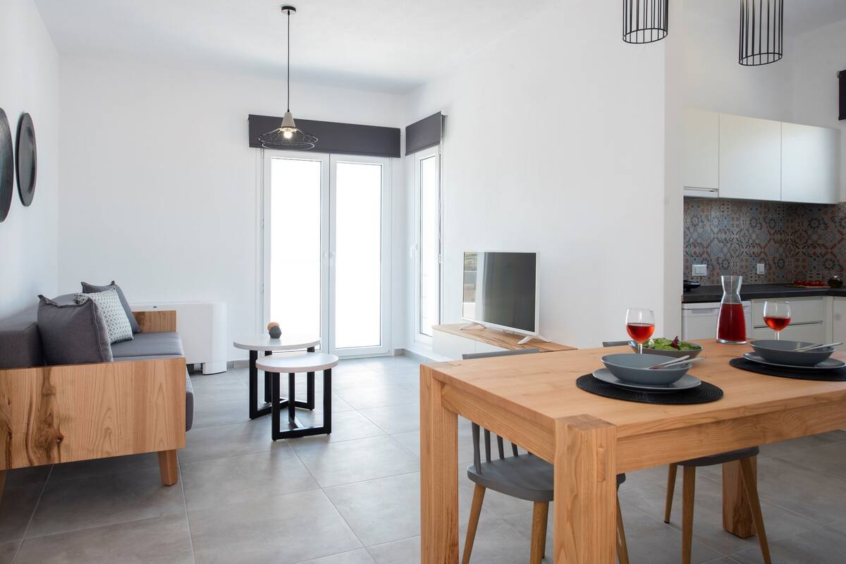 Una sala moderna con un sofá de madera, una mesa de comedor con dos sillas, un televisor y una zona de cocina con armarios y una encimera.
