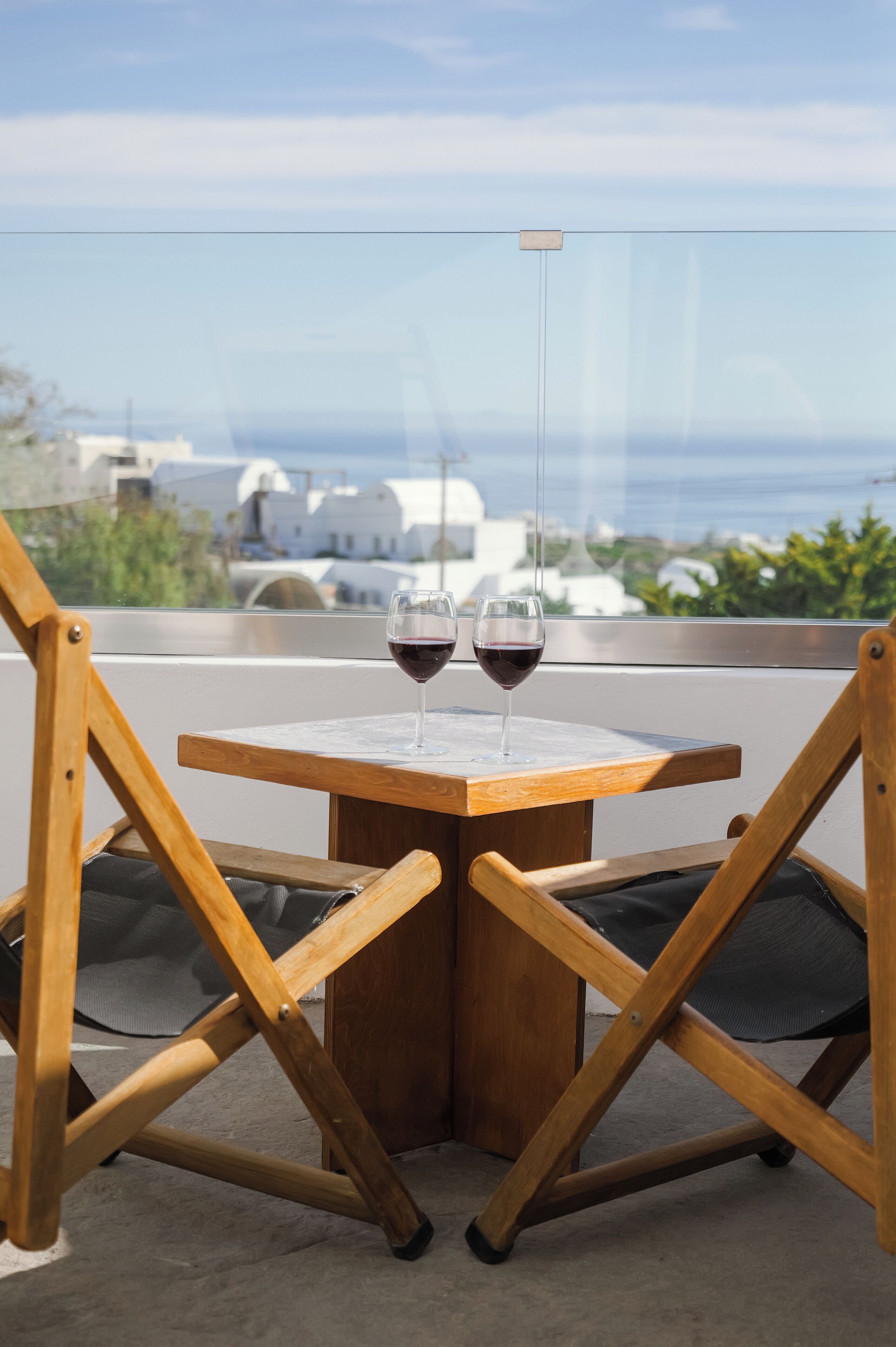 Una mesa de madera con dos copas de vino tinto, vista al mar y edificios blancos al fondo.