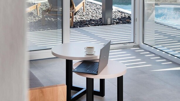 Una habitación moderna con una computadora portátil sobre una mesa redonda, ventanas amplias con vista a una piscina y paisaje, y una lámpara colgante.