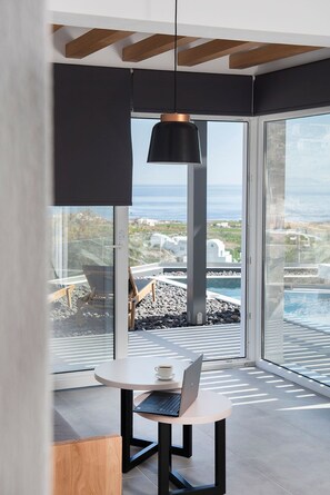 A modern interior with a laptop on a table, a hanging light fixture, and a view of the sea through large windows. - Valery Suites (Santorini)