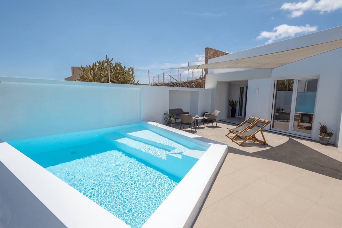 Un área de piscina moderna al aire libre con una piscina azul cristalina, un espacio de descanso con sillas y una mesa, y una puerta de cristal que da a un porche cubierto.