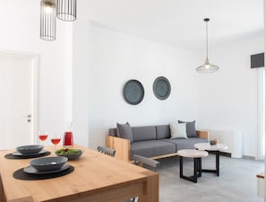 A modern living room with a grey sofa, a wooden dining table with bowls and glasses, and a small round table with a vase. - Valery Suites (Santorini)