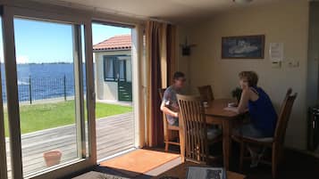 A room with a large sliding glass door leading to a deck with a view of the water. There is a dining table with chairs, a small potted plant, and a remote control on the table.