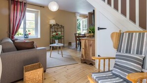 A cozy living room with a sofa, a dining table with chairs, a staircase, and a window with curtains. - Charming house in the valley for 2, nature and tranquility (Beauficel)