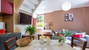 A dining area with a table set for two, a television mounted on the wall, and a sofa in the background. - Charming house in the valley for 2, nature and tranquility (Beauficel)