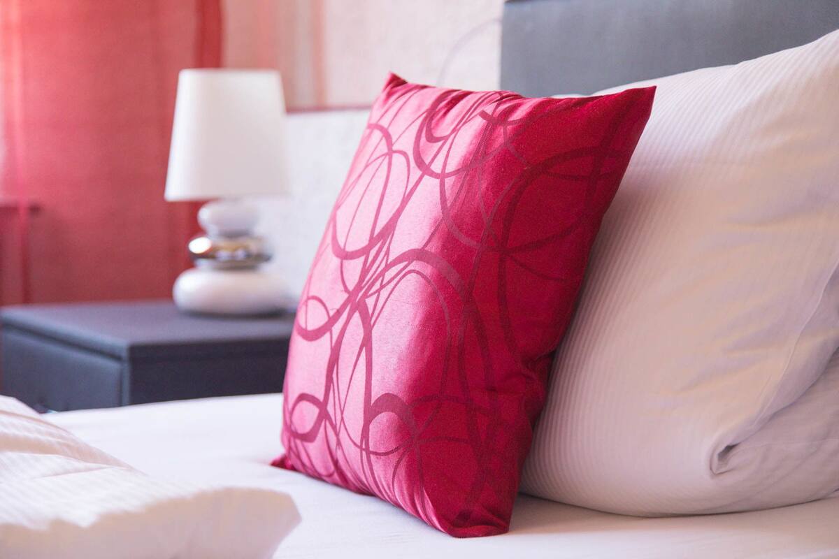 A bed with a red and white pillow, a white lamp on a bedside table, and a red curtain in the background.