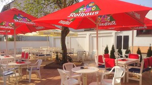 An outdoor patio area with white plastic chairs and tables, red umbrellas with 'Rothaus' branding, and a tree in the center. - Hotel Berlin (Schwetzingen)