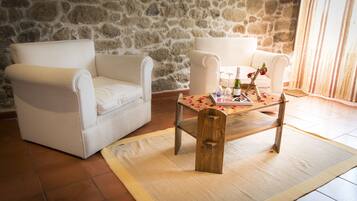 Quarto com parede de pedra, duas poltronas brancas, uma mesa de centro de madeira com um descanso para prato, e piso ladrilhado.