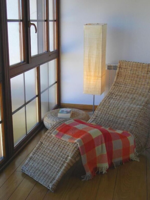 A room with a large window, a woven chair, a small round table, and a tall floor lamp.