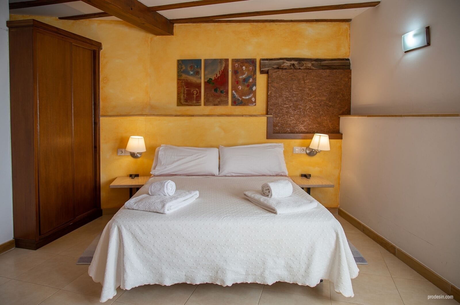 A hotel room with a large bed, two bedside lamps, a wooden wardrobe, and artwork on the walls.