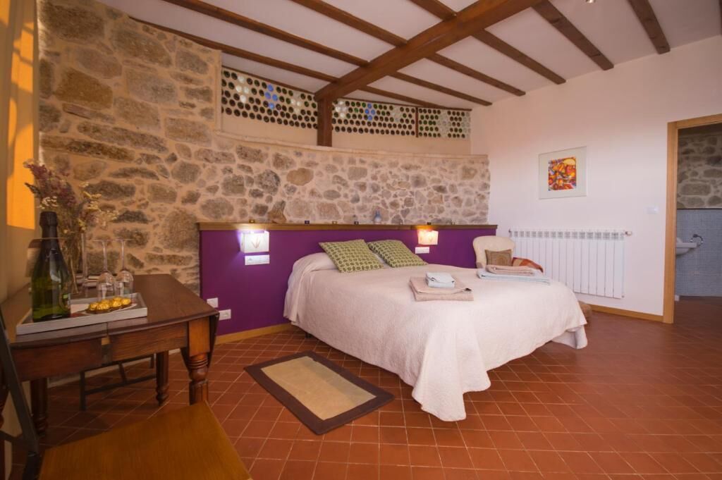 A bedroom with a stone wall, a bed with a white bedspread, a wooden table with a bottle and glasses, and a window with a view of the outside.