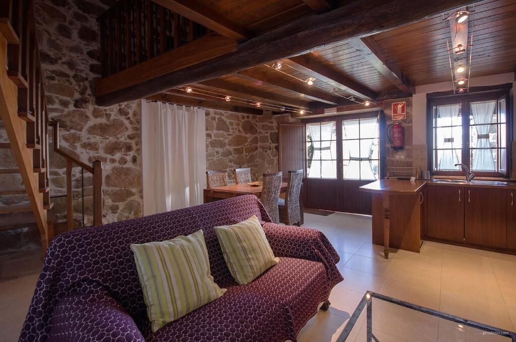 Un interior acogedor con pared de piedra, vigas de madera, un sofá morado estampado, una mesa de comedor y una zona de cocina.