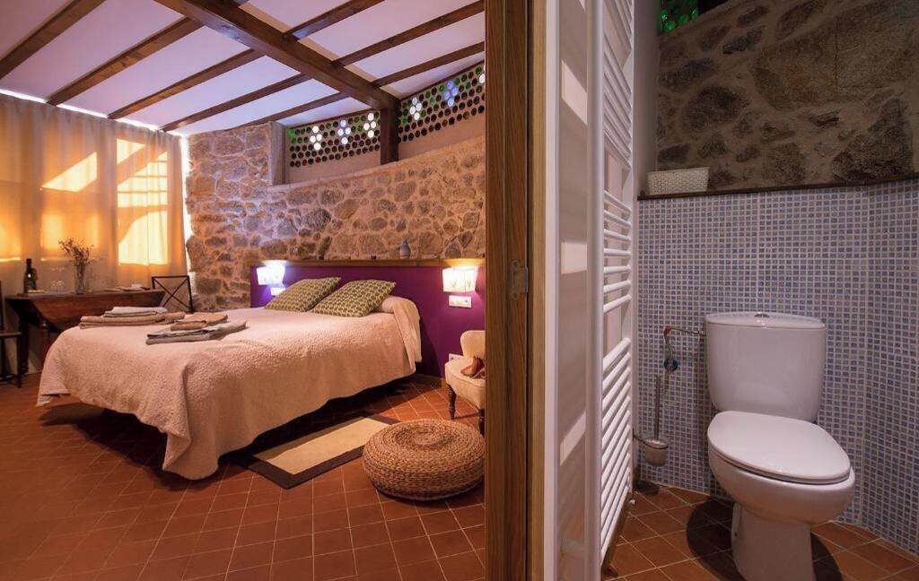 A bedroom with a stone wall, a bed with a headboard, a bedside table, and a bathroom with a toilet.