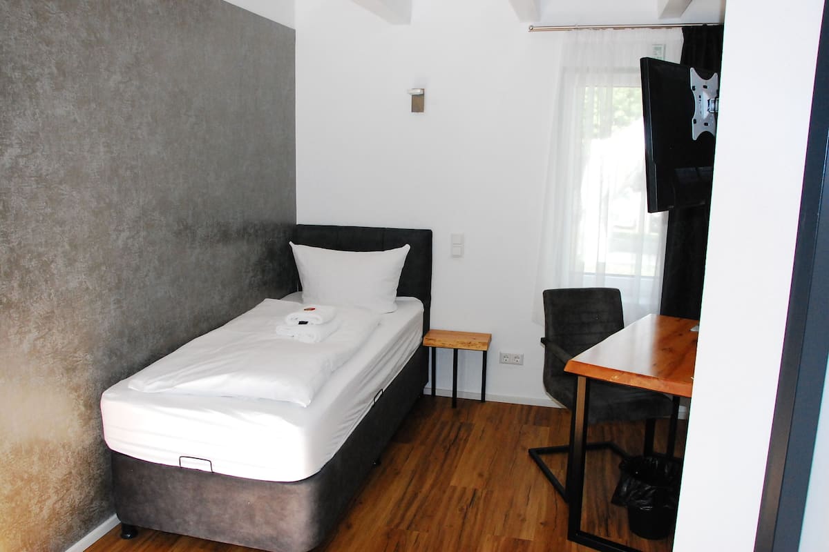 Ein kleines Zimmer mit einem Einzelbett, einem kleinen Tisch, einem Stuhl und einem an der Wand befestigten Fernseher.
