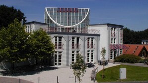 A modern building with a curved glass facade, a flat roof, and a balcony with red railings. - TaT Hotel (Rheine)