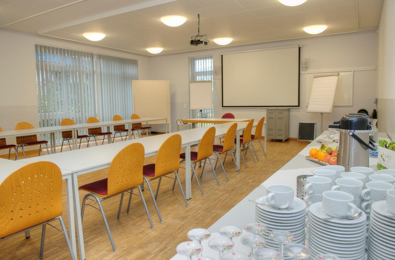 A conference room with long tables, chairs, a projector screen, and a buffet area with plates and cups.