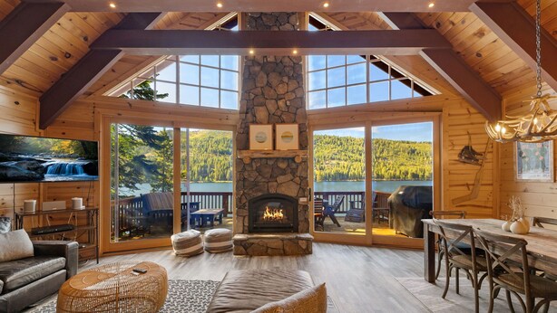 Un intérieur de chalet chaleureux avec une cheminée en pierre, des poutres en bois, de grandes fenêtres et un coin repas.