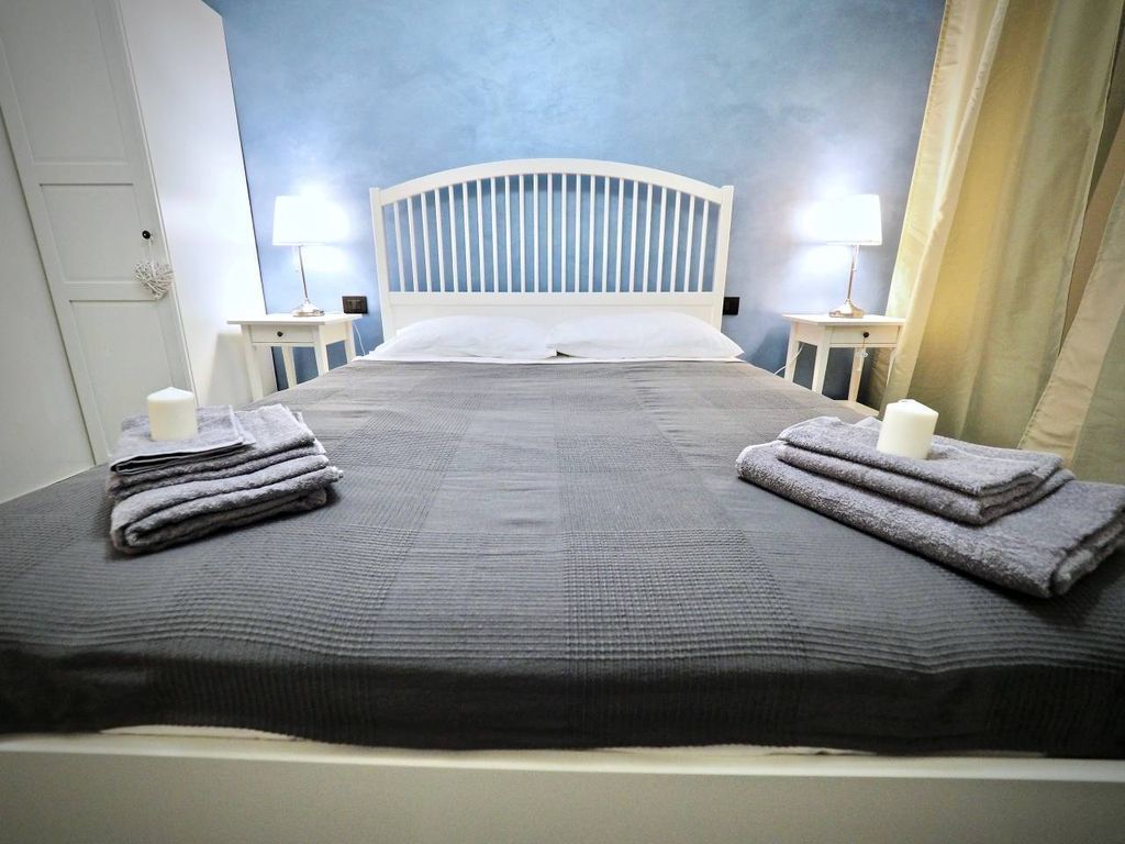 Een net opgemaakt bed met een grijze dekbedovertrek en twee gevouwen handdoeken aan elke kant. Er zijn twee nachtkastjes met lampen, één aan elke zijde van het bed.