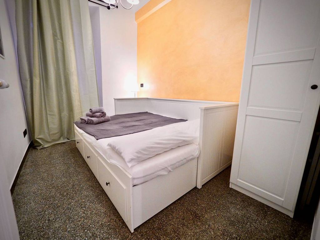 A small, single-bed room with a white bed, a grey blanket, and a towel on the bed. There is a white wardrobe to the right and a door to the left.