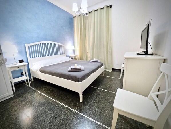 Ein Schlafzimmer mit Bett, Schreibtisch, Stuhl, Fernseher und einem Fenster mit Vorhängen.