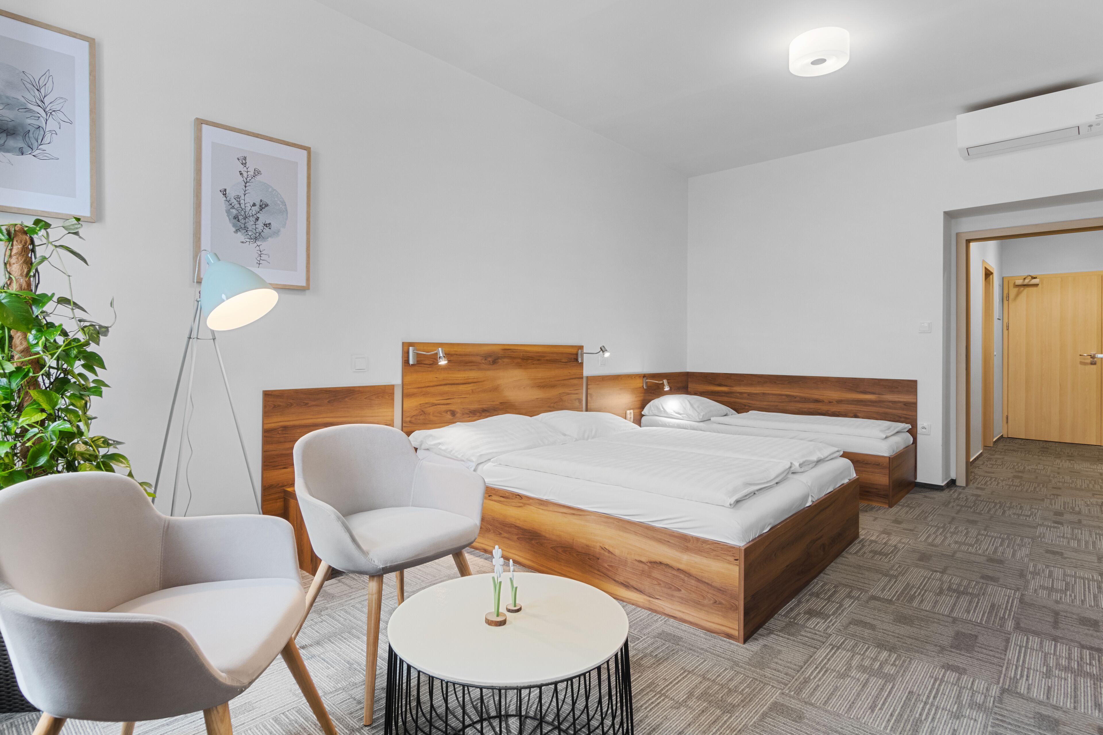 Una habitación de hotel moderna con una cama con cabecera de madera, dos sillas blancas, una mesita redonda y una planta en maceta.
