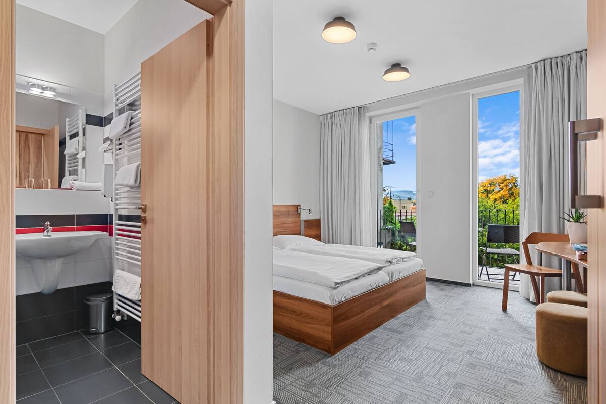 Ein modernes Hotelzimmer mit einem großen Bett, einem separaten Badezimmer mit Waschbecken und Handtuchhalter, einem kleinen Tisch und Blick ins Freie.