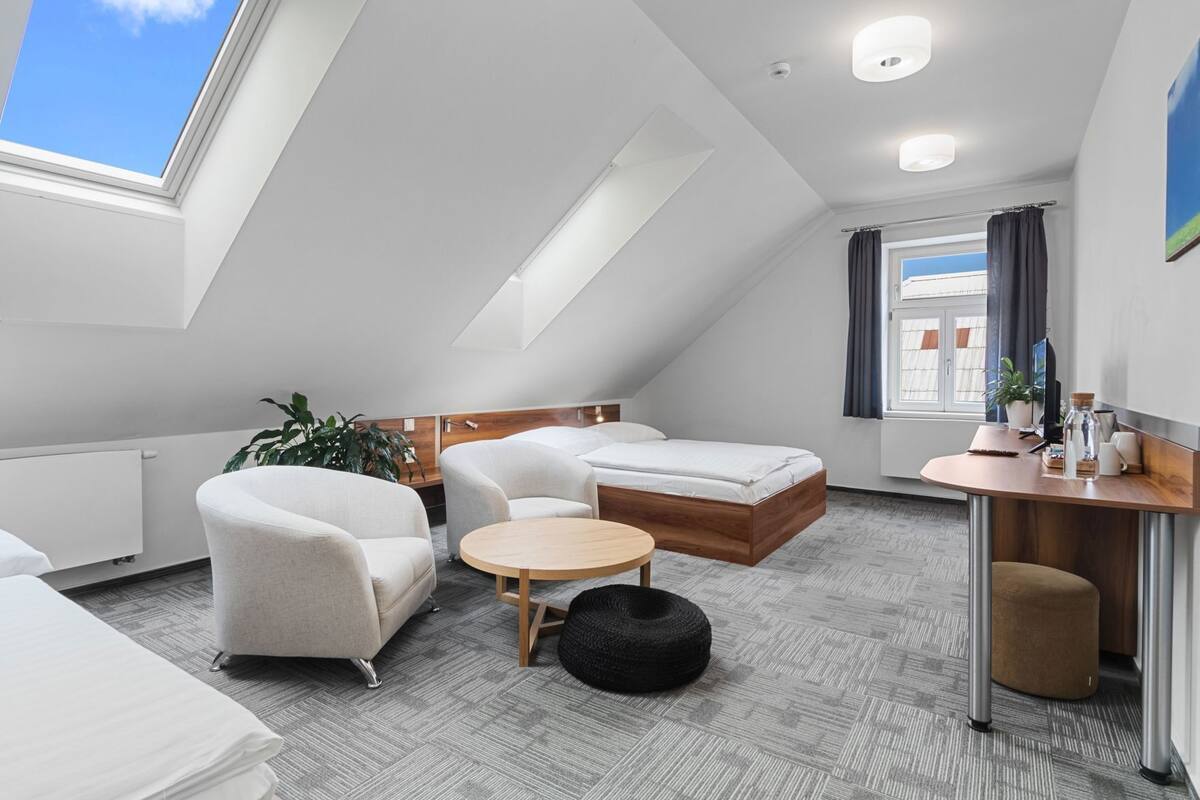 Ein modernes Hotelzimmer mit schrägverdeckter Decke, einem Bett, einem Sofa, einem kleinen runden Tisch, einem Schreibtisch mit Stuhl, einer Topfpflanze und einem Fenster mit Vorhängen.
