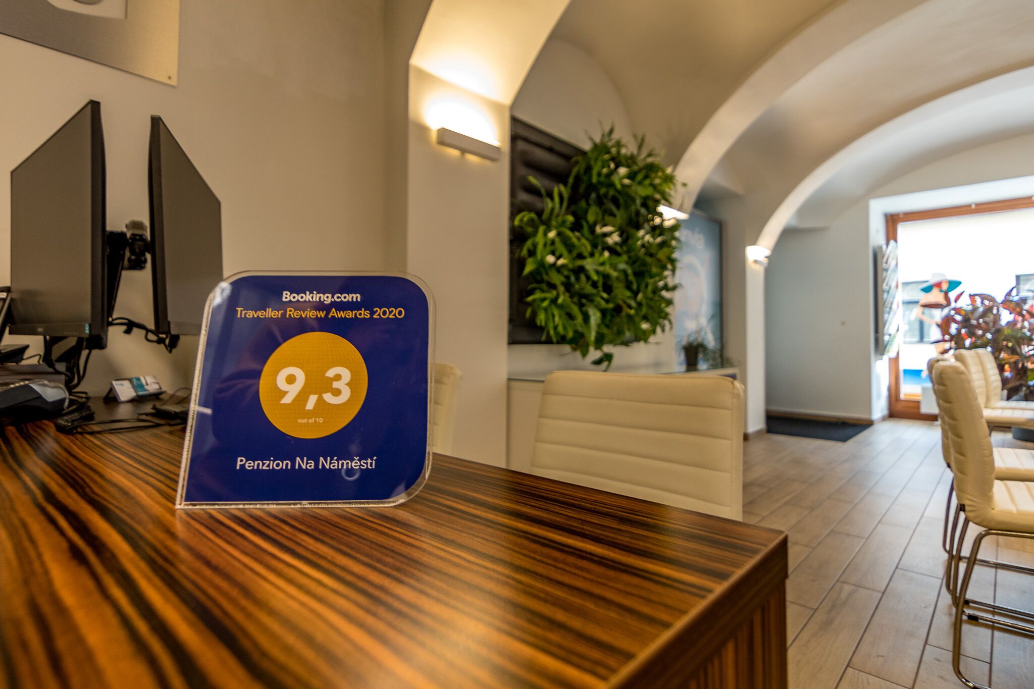 Una recepción con una placa azul de reconocimiento de Booking.com, una planta en maceta y vista al vestíbulo.