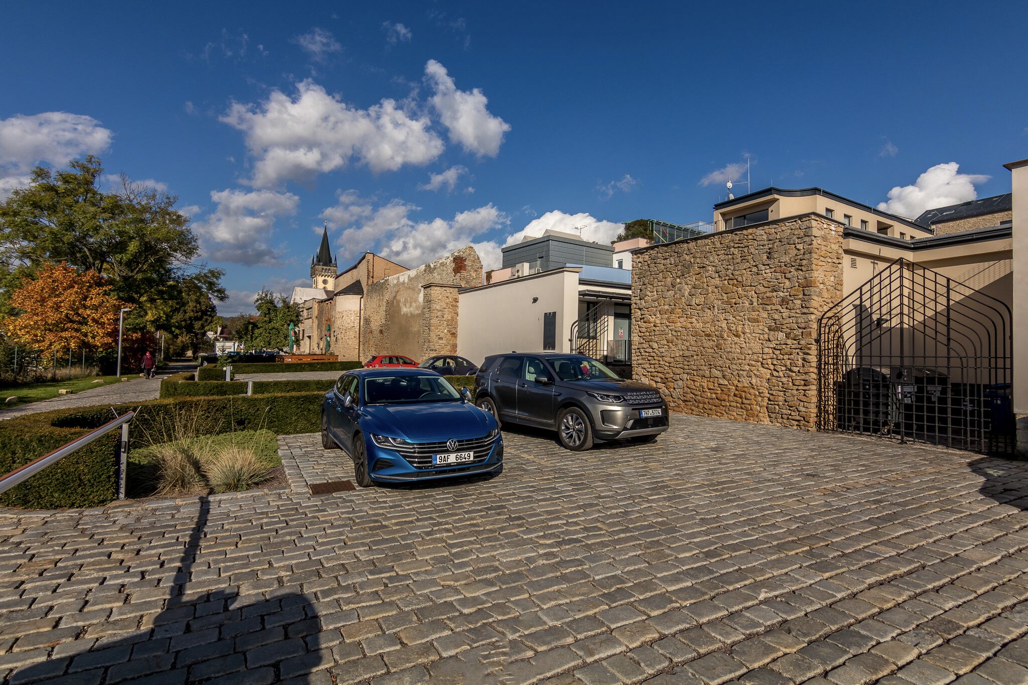 Ein Parkplatz mit zwei Autos, einem Steinhaus und einer Kirche im Hintergrund.