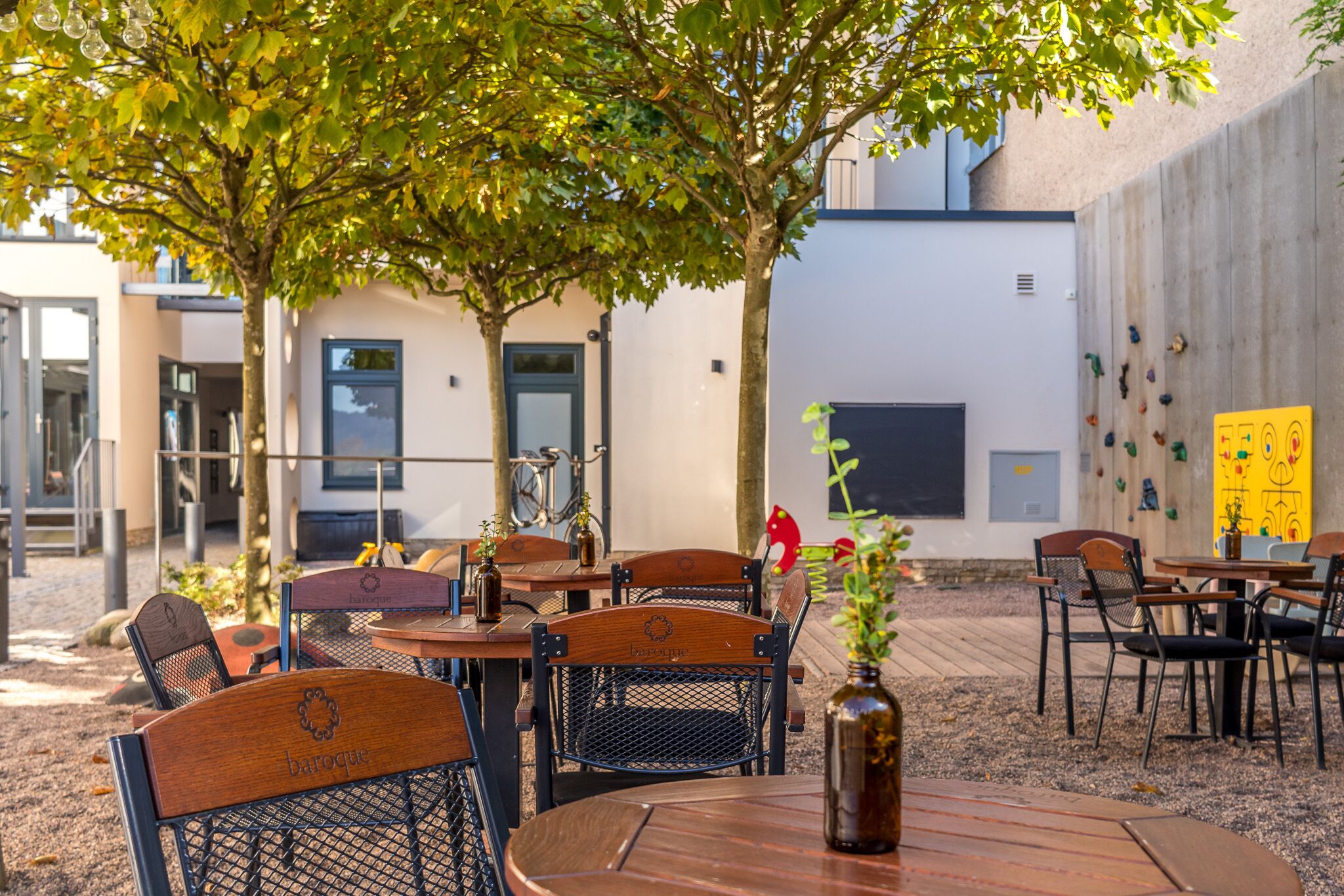 An outdoor seating area with wooden tables and chairs, a decorative vase, and a climbing wall in the background.