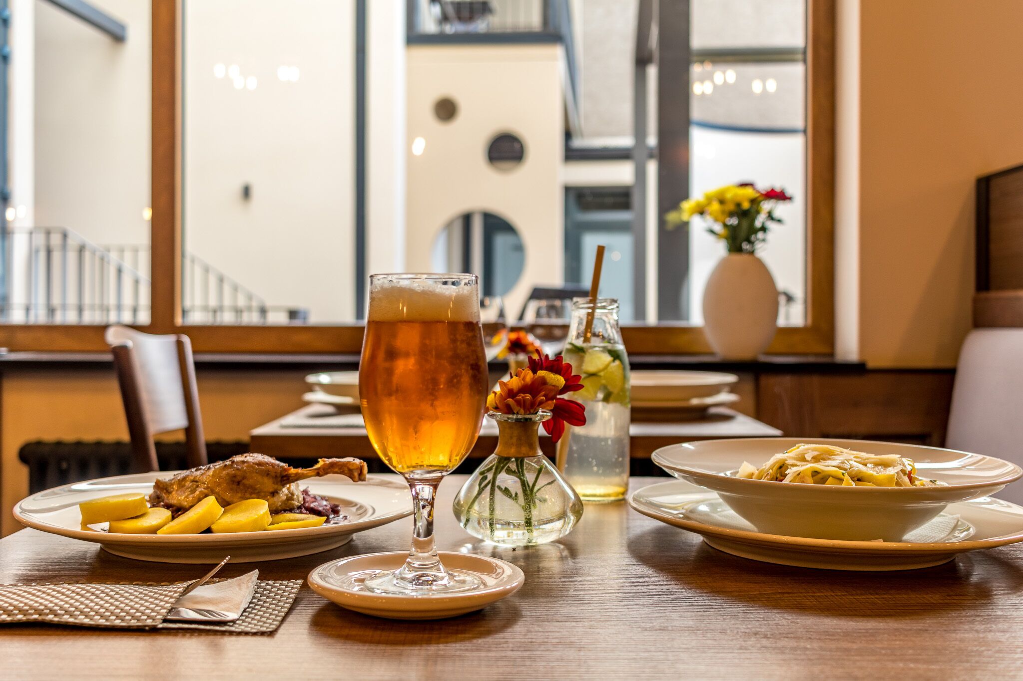 Une table à manger avec des assiettes remplies de nourriture, un verre de bière et un vase de fleurs.